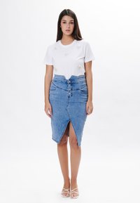 Camiseta blanca con detalles bordados, combinada con una falda lápiz de mezclilla azul claro de cintura alta, que presenta una abertura en el frente y cierre de botones.