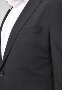 Zwarte colbert van een gestructureerde stof, met een enkele knoopsluiting en een notch revers, voorzien van een borstzak en een wit overhemd eronder.