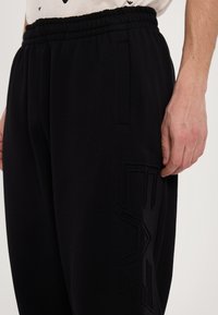 Black sweatpants with an elastic waistband and side pockets; features a textured logo detail on the left leg. Soft fabric with a smooth finish.