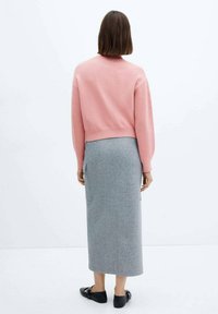 Femme debout de dos, portant un pull rose, une jupe midi grise avec poche latérale et des chaussures plates noires sur un fond uni.