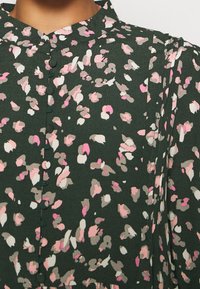 Blouse vert foncé avec un motif floral abstrait rose, blanc et beige, et des boutons recouverts de tissu sur le devant.