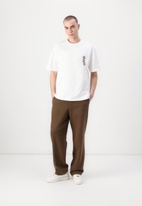 White cotton t-shirt with a small dark floral print on the left chest, paired with brown trousers and white sneakers. Simple and relaxed fit.
