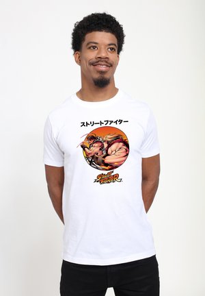 Mand iført hvid T-shirt med Street Fighter-grafik, der viser to muskuløse kæmpere i kamp og japansk tekst over designet.