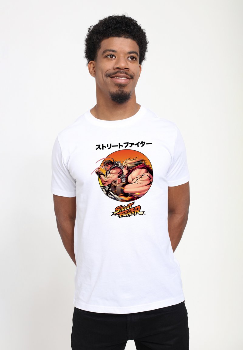 Homme portant un T-shirt blanc avec un graphisme Street Fighter montrant deux combattants musclés en pleine bataille et du texte japonais au-dessus du design.