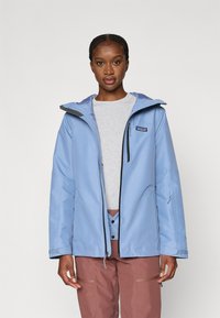 Αδιάβροχο ανοιχτό γαλάζιο μπουφάν με κουκούλα, μαύρο φερμουάρ και λογότυπο στο πάνω αριστερό μέρος του στήθους. Φορεμένο πάνω από γκρι πουκάμισο και καφέ παντελόνι.