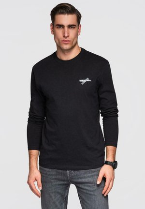 Jeune homme avec les cheveux soigneusement coiffés portant une chemise noire à manches longues avec un petit logo blanc sur la poitrine et un jean gris.