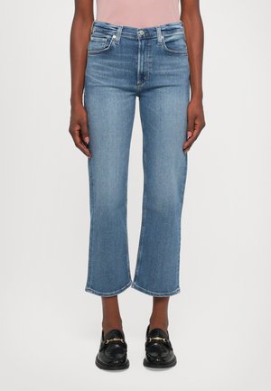 Mujer vestida con jeans cortos azules y mocasines de cuero negros con cadenas doradas, de pie frente a un fondo blanco liso.