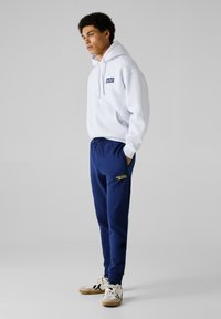 Weißer Hoodie mit Logo auf der Vorderseite, marineblaue Jogginghose mit gelber Schrift und weiße Sneakers mit schwarzen und sandfarbenen Akzenten. Einfaches sportliches Design.