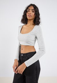Fiatal nő, göndör hajjal, világosszürke, hosszú ujjú crop toppot és magasderékú fekete farmert visel, egy egyszínű háttér előtt pózol.