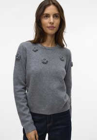 Frau in einem grauen Wollpullover mit kleinen gestrickten Blumenapplikationen, kombiniert mit dunkelblauen Jeans, vor einem weißen Hintergrund.