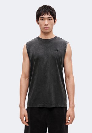 Junger Mann, der ein verblichenes schwarzes ärmelloses Shirt und schwarze Hose trägt, steht frontal vor einem einfarbigen Hintergrund.