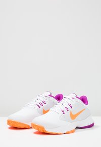 Białe buty sportowe z fioletowymi akcentami i pomarańczowym logo Nike. Posiadają siateczkowy wierzch, teksturowaną podeszwę oraz sznurowane zapięcie.