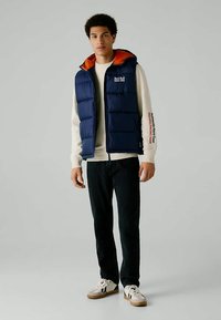 Νεαρός άνδρας φορώντας ένα ναυτικό μπλε επενδυμένο γιλέκο Red Bull Racing πάνω από έναν κρεμ φούτερ, μαύρο παντελόνι και λευκά αθλητικά παπούτσια, στέκεται σε γκρι φόντο.