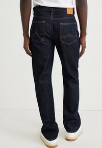 Man som bär mörkblå raka jeans med kontrastsömmar och vita sneakers, stående mot en helt ljus bakgrund.