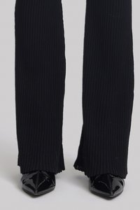 Pantalons noirs côtelés à coupe évasée, avec des ourlets effilochés, associés à des talons aiguilles en cuir verni noir. Texture lisse et design épuré.