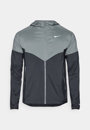Chaqueta con capucha de Nike que presenta un diseño degradado con la parte superior en gris claro y la parte inferior en gris oscuro. Fabricada con un tejido suave y ligero, con cierre de cremallera.