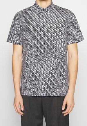 Man wearing short-sleeve button-up shirt with all-over black "HUGO" text pattern on white background and dark trousers.