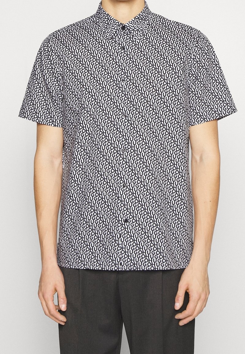 Homme portant une chemise à manches courtes à boutons avec un motif intégral "HUGO" noir sur fond blanc et un pantalon sombre.