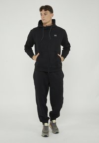 Haut à fermeture éclair noir avec cordons de serrage, présentant un petit logo sur la poitrine, associé à un pantalon de jogging noir assorti et des chaussures de sport grises.