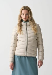 Puffjacka i ljusbeige med quiltad design, hög krage och framdragkedja. Slät textur med elastiska ärmslut. Bärs över en teal-färgad kjol.