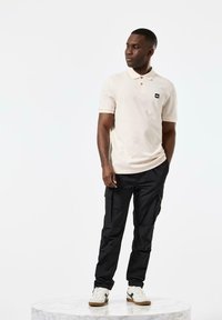 Polo skjorte i lys beige bomull, med to knapper og en liten svart logo. Kombinert med svarte cargo-bukser og hvite sneakers med brune detaljer.