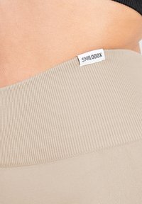 Beige geribbelde tailleband met een klein wit "SMILODOX" label, geïntegreerd in het ontwerp van een aansluitend kledingstuk. Zachte, gladde textuur.