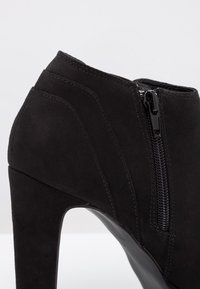 Bottines à talons hauts en daim noir avec fermeture éclair latérale et design de panneaux courbes. Texture lisse avec un accent de fermeture éclair brillante.