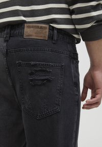 Jeans in denim nero con una tasca posteriore usurata e un'etichetta in pelle marrone con il logo del brand. Il tessuto sembra resistente e ha una consistenza morbida.