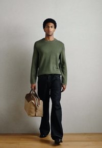 Camisola verde de malha, jeans escuros e sapatos pretos, a carregar uma mala castanha com detalhes em marrom. Design simples e funcional, padrões mínimos.