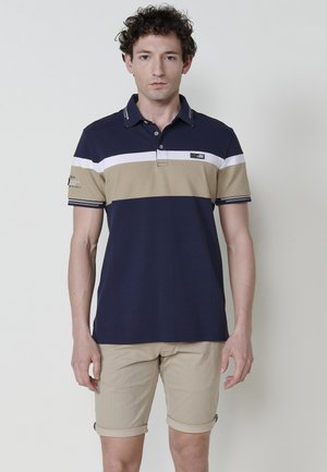 Camisa polo azul marino con rayas horizontales beige y blancas, de mangas cortas y con cuello. Llevada con pantalones cortos beige claros enrollados.