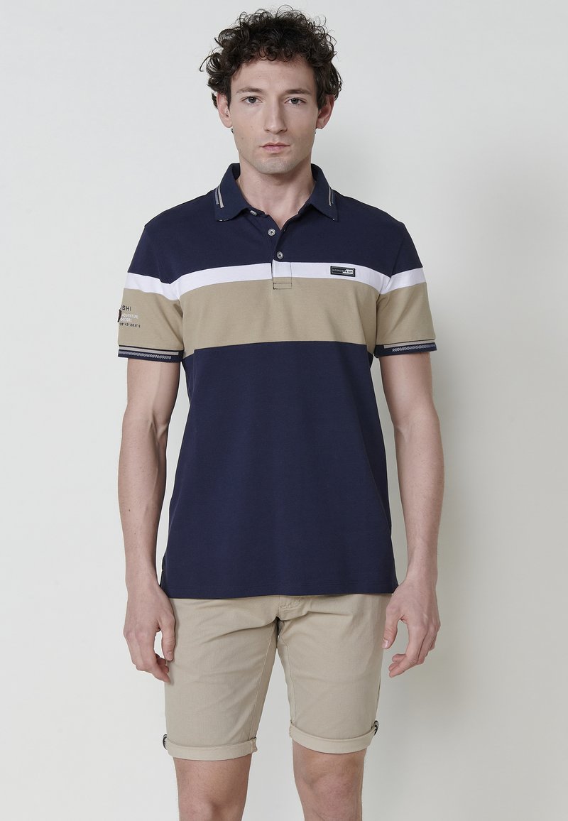 Sötétkék póló ing bézs és fehér vízszintes csíkokkal, rövid ujjakkal és gallérral. Világos bézs, feltekert rövidnadrággal viselve.