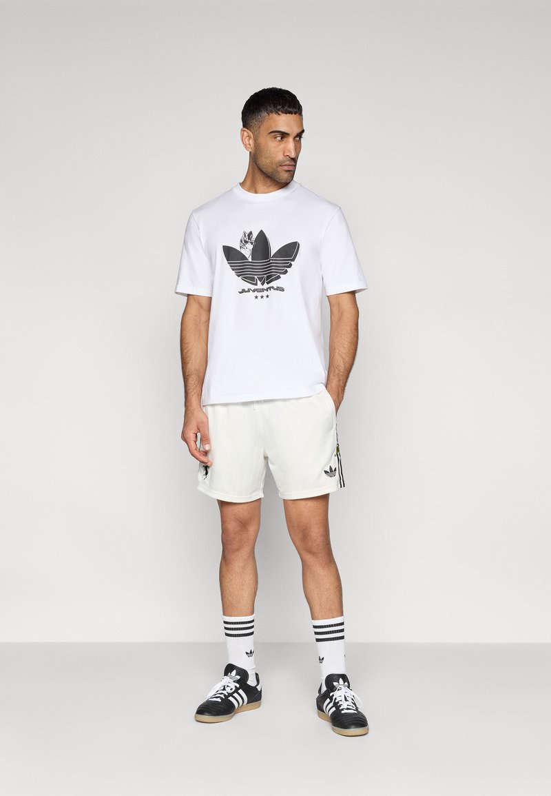 Weißes T-Shirt mit schwarzem Adidas-Logo, kombiniert mit weißen Shorts und schwarzen Sneakern. Mit gestreiften Socken und minimalistischem Design.