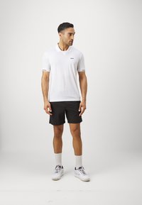 Λευκό κοντομάνικο πόλο μπλουζάκι με μαύρο λογότυπο, συνδυασμένο με �μαύρη βερμούδα. Το μοντέλο φοράει λευκά αθλητικά παπούτσια και κάλτσες μέτριας μήκους. Απλός σχεδιασμός.