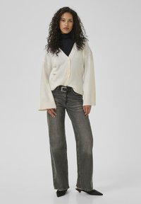 Cream-colored oversized cardigan with buttons, paired with dark high-waisted jeans. Worn over a black turtleneck and black pointed shoes.