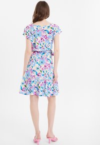 Blumenmusterkleid mit blauem, pinkem und gelbem Muster; kurze Ärmel, geraffter Saum und taillierter Schnitt; kombiniert mit pinken High Heels.