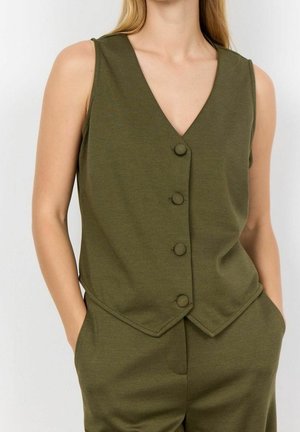 Frau trägt ärmellose, olivgrüne Weste mit Knöpfen und V-Ausschnitt sowie passende Hose, Hände in den Taschen, vor einfarbigem Hintergrund.