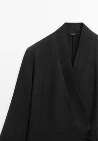 Blazer noir sur mesure avec larges revers et devant croisé, confectionné en tissu texturé, présenté sur un fond blanc.