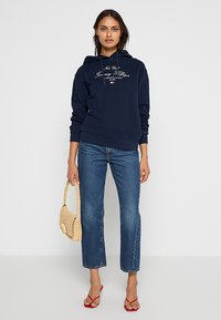 Felpa blu navy con cappuccio regolabile e testo bianco, abbinata a jeans blu a gamba dritta e sandali rossi, mentre tiene una borsa di paglia beige.