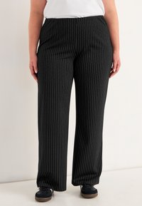 Persoon in zwarte broek met dunne witte verticale streepjes, zwarte sneakers met witte zolen, en een wit T-shirt, staand tegen een lichte achtergrond.