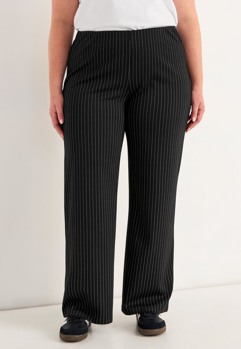 Persoon in zwarte broek met dunne witte verticale streepjes, zwarte sneakers met witte zolen, en een wit T-shirt, staand tegen een lichte achtergrond.