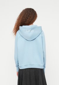 Άτομο με σγουρά μαλλιά που φοράει ένα ανοιχτό μπλε φούτερ με κουκούλα, πάνω στην οποία υπάρχει το κείμενο "LeGer", συνδυασμένο με μια σκούρα πλισέ φούστα, με την πλάτη να κοιτάζει.