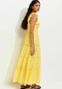 Mujer con cabello largo y oscuro que lleva un vestido maxi amarillo sin mangas de capas con tirantes con volantes y sandalias marrones, parada contra una pared blanca.
