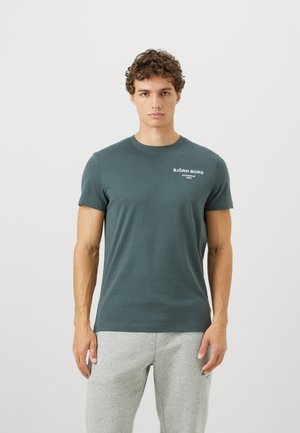 Ung mand med krøllet hår iført en mørkegrøn Björn Borg t-shirt og lysegrå sweatpants, stående foran en ensfarvet hvid baggrund.