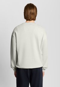 Άτομο με ανοιχτά καστανά μαλλιά, που φοράει μονόχρωμο ανοιχτό γκρι φούτερ και σκούρο παντελόνι, γυρισμένο με την πλάτη προς το φακό, μπροστά σε απλό γκρι φόντο.