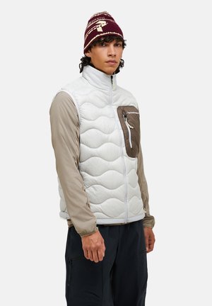 Ung mand iført en hvid quiltet vest med en brun lynlåslomme på brystet, beige lange ærmer, sorte bukser og en mørkerød beanie.