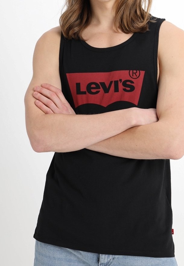 Personne portant un débardeur noir Levi's, les bras croisés, avec un jean bleu clair sur un fond blanc.
