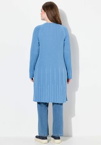 Hellblauer langer Cardigan mit vertikaler Rippstruktur, abgerundetem Saum und seitlichen Schlitzen, kombiniert mit hellblauen Jeans und lässigen dunkel gefärbten Schuhen.