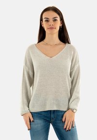 Jersey de punto gris claro con cuello en V y mangas holgadas, que presenta un patrón texturizado y un corte relajado, combinado con vaqueros azules.