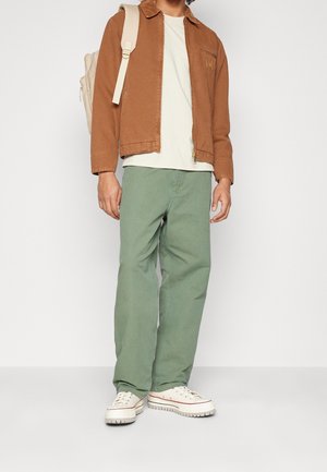 Mężczyzna w brązowej kurtce, białej koszulce, zielonych spodniach, białych trampkach, niosący beżowy plecak, stoi na białym tle.