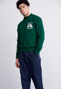 Groene sweatshirt met ronde hals en geribbelde manchetten, voorzien van een witte grafische opdruk van "Ellesse 1959" en "Club di Corsa" op de borst. Gecombineerd met donkere marineblauwe broek.